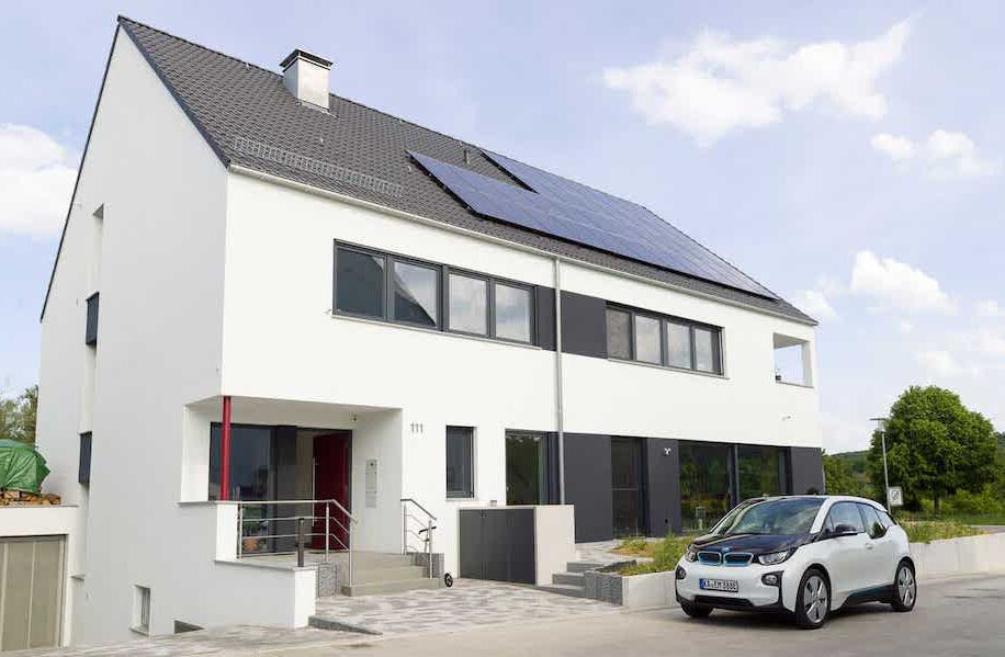 Blick auf ein freistehendes Einfamilienhaus an einer Strasse mit parkendem Auto davor. Das Haus ist weiß gestrichen, zweigeschossig und hat ein Satteldach, auch welchem sich eine PV Anlage befindet.