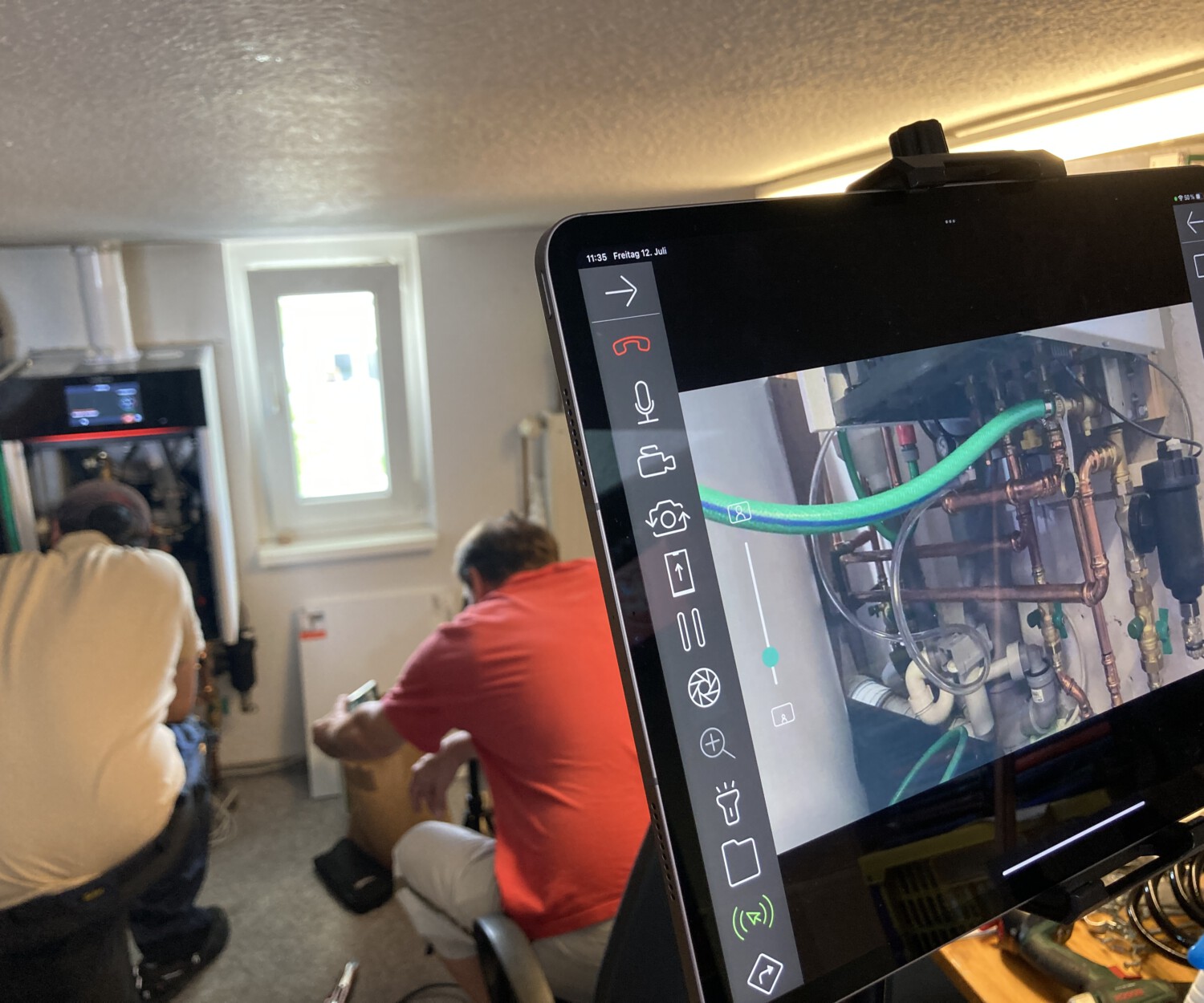 Tablett PC auf Stativ, auf dem Tablett PC ist das Live Bild aus dem Inneren einer Heizungsanlage abgebildet, die Bauteile sind mit Overlay Pfeilen beschriftet.